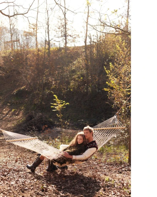A young couple is relaxing in a hammock in a sunny wooded setting, wearing sweaters and boots; low-angle view.