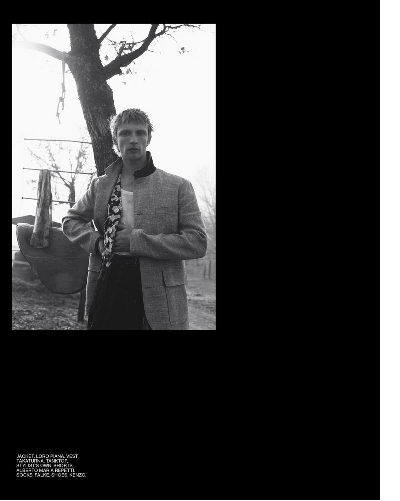 Black and white portrait of a blonde male model wearing a jacket and patterned shirt outdoors with a tree and nature background.