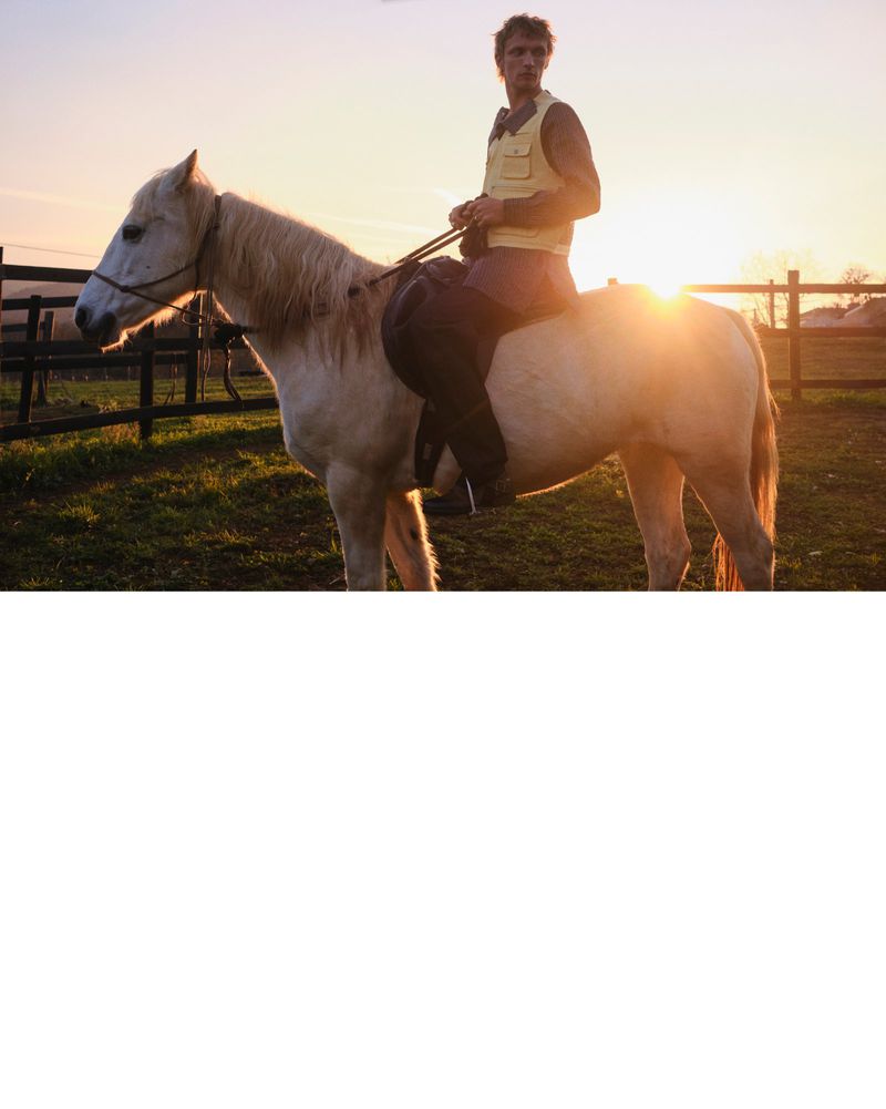 A rider on a white horse stands in a sun-drenched pasture with a wooden fence, illuminated by golden light, capturing a rural scene.