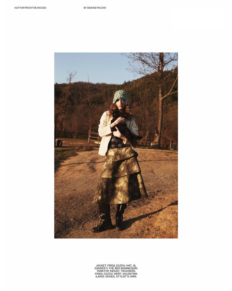 A young woman in a patterned hat and skirt poses in a woodland setting, exuding a blend of nature-inspired fashion and serene outdoor ambiance.