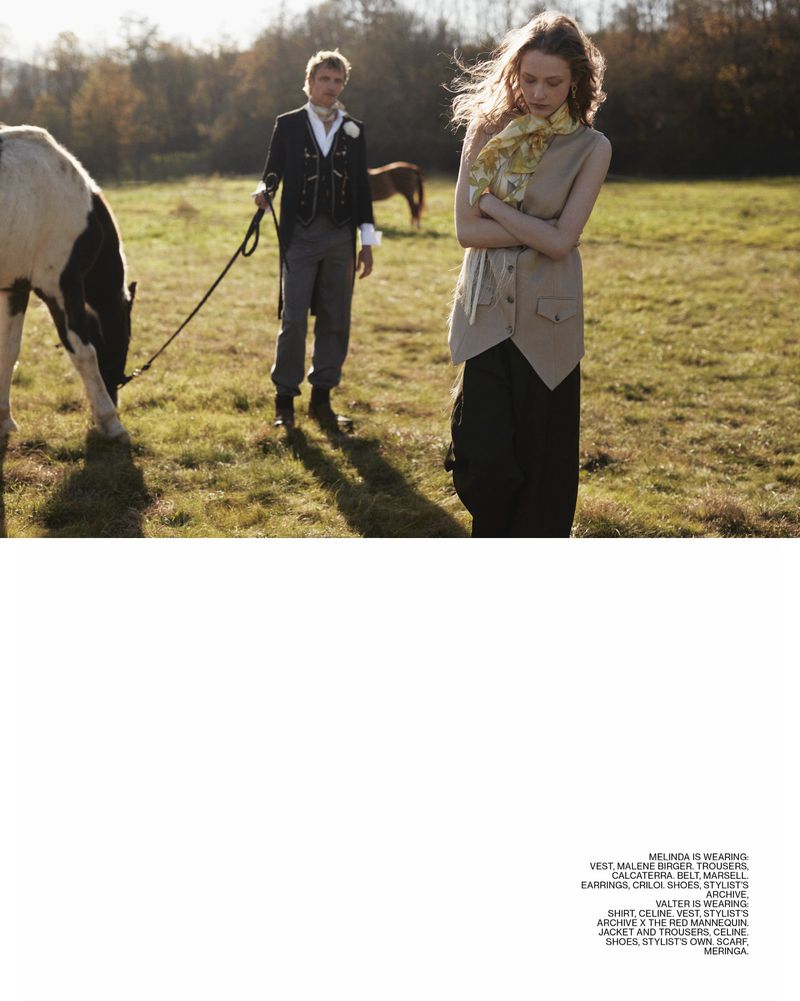 Fashion editorial image in grassy field. A woman in a vest stands with arms crossed as a man walks a horse in the background.