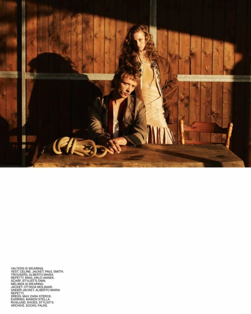 Two fashion models pose in front of a wooden wall with natural light casting shadows, wearing stylish clothing and neutral colors.