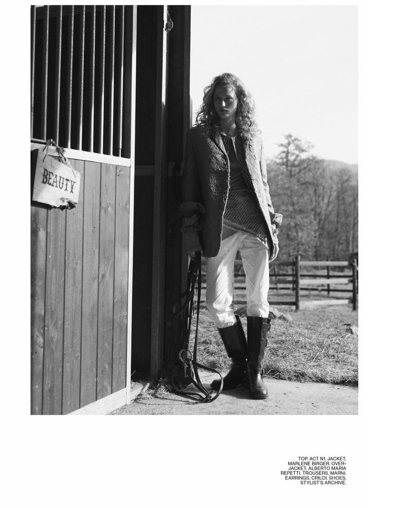 A slender woman with curly hair stands in a stable wearing a blazer, top, white jeans, and tall boots, holding reins in a black-and-white fashion shot.