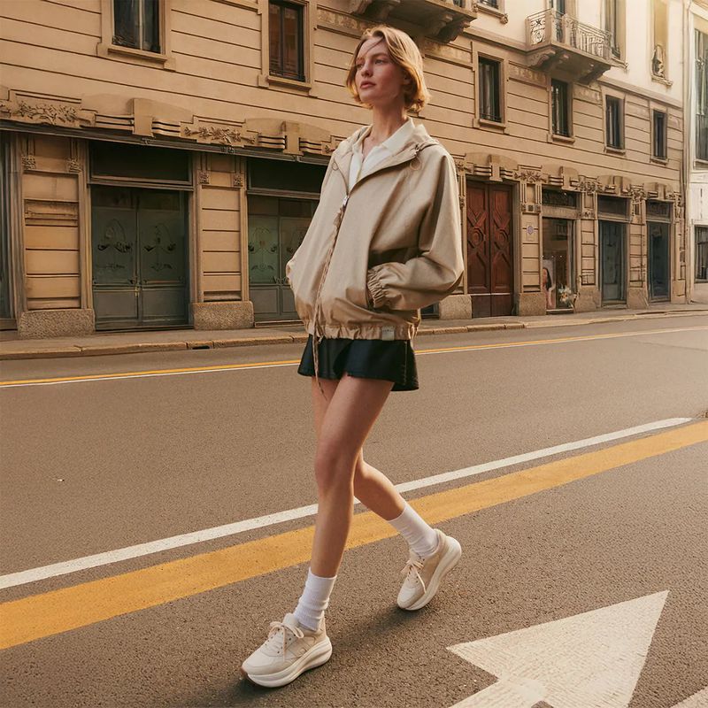 Woman in beige jacket and black skirt walks across a city street with building facades and warm lighting.