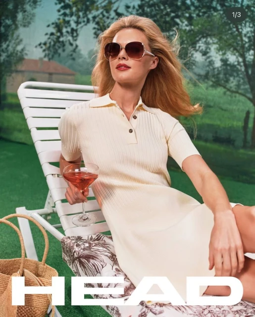 A blonde woman in sunglasses wearing a cream-colored dress sits on a white lawn chair holding a drink in a lush outdoor setting.