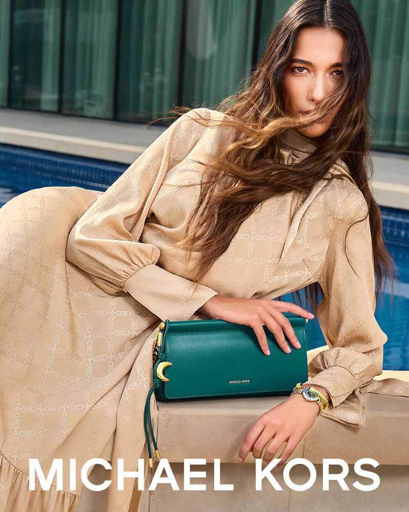 A woman in a beige dress poses with a green Michael Kors purse in an outdoor setting with natural light and a modern aesthetic.