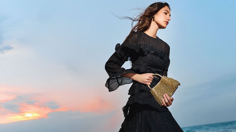 Woman in ruffled black dress holding a beaded gold handbag stands near a sunset on a beach with the ocean visible in the distance.
