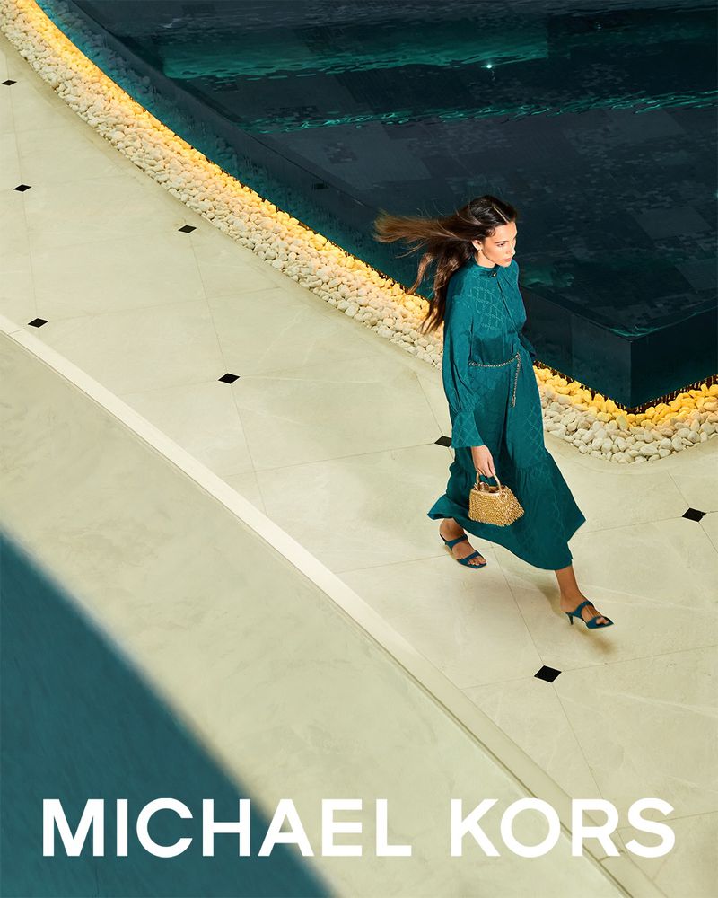 Model in a long teal dress walking by a swimming pool holding a woven handbag for Michael Kors Ramadan campaign