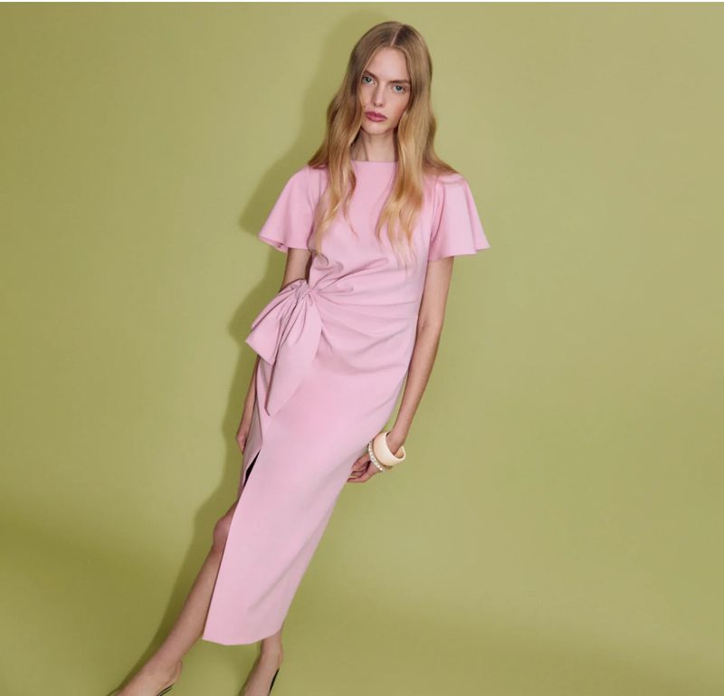 Elegant woman in a pastel pink midi dress with a side bow posing against a pale green background in a fashion-forward studio shot.