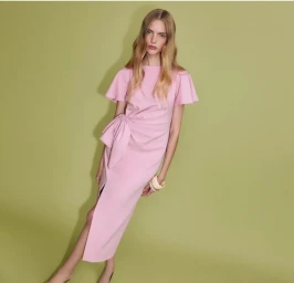Model Eduarda Bretas in a pink dress with side bow posing against a green backdrop for Tanya Taylor Spring 2026