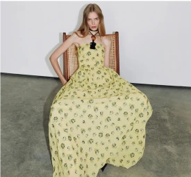 Model seated in a cane-backed chair wearing a yellow floral maxi dress for Tanya Taylor Spring 2026.