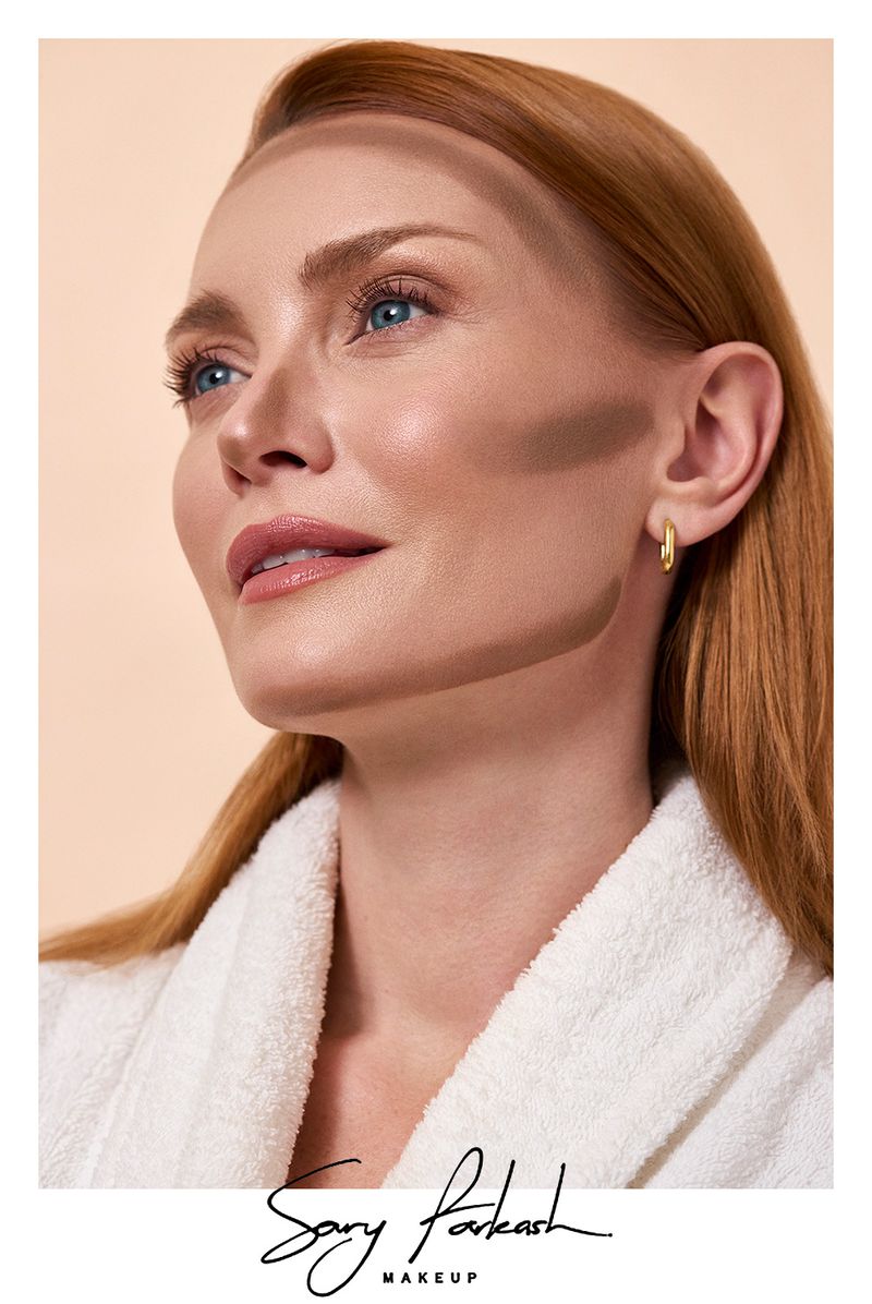 Close-up portrait of a fair-skinned woman with auburn hair wearing a white robe and gold earrings. Face is contoured with brown makeup.