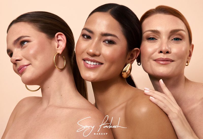 Three diverse women pose together with gold earrings against a tan backdrop for a beauty campaign promoting makeup.