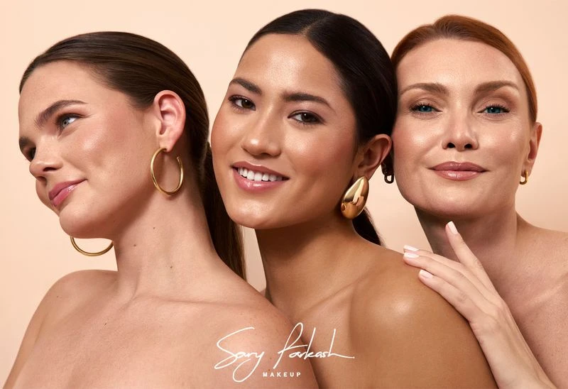 Three diverse women pose together with gold earrings against a tan backdrop for a beauty campaign promoting makeup.