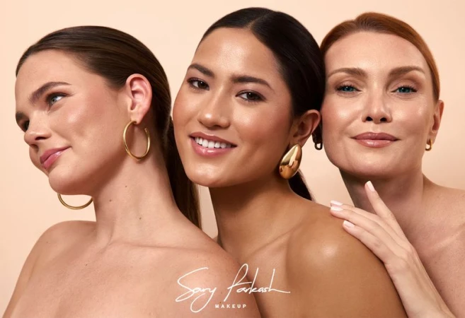 Three diverse women pose together with gold earrings against a tan backdrop for a beauty campaign promoting makeup.