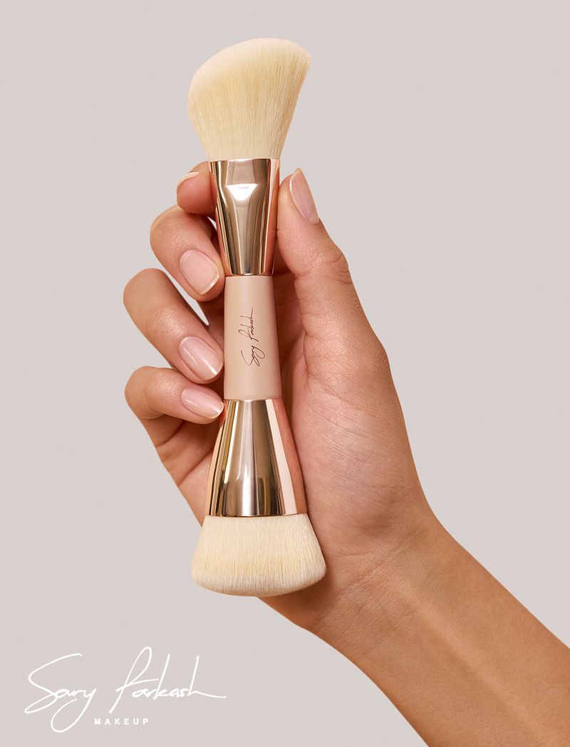 Tan-skinned hand holds a double-ended makeup brush with rose gold accents against a neutral background in a beauty-focused close-up shot.