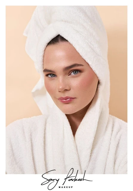 Close-up portrait of a fair-skinned woman in a white bathrobe and towel wrap, featuring a peach-toned makeup palette with glossy lips.