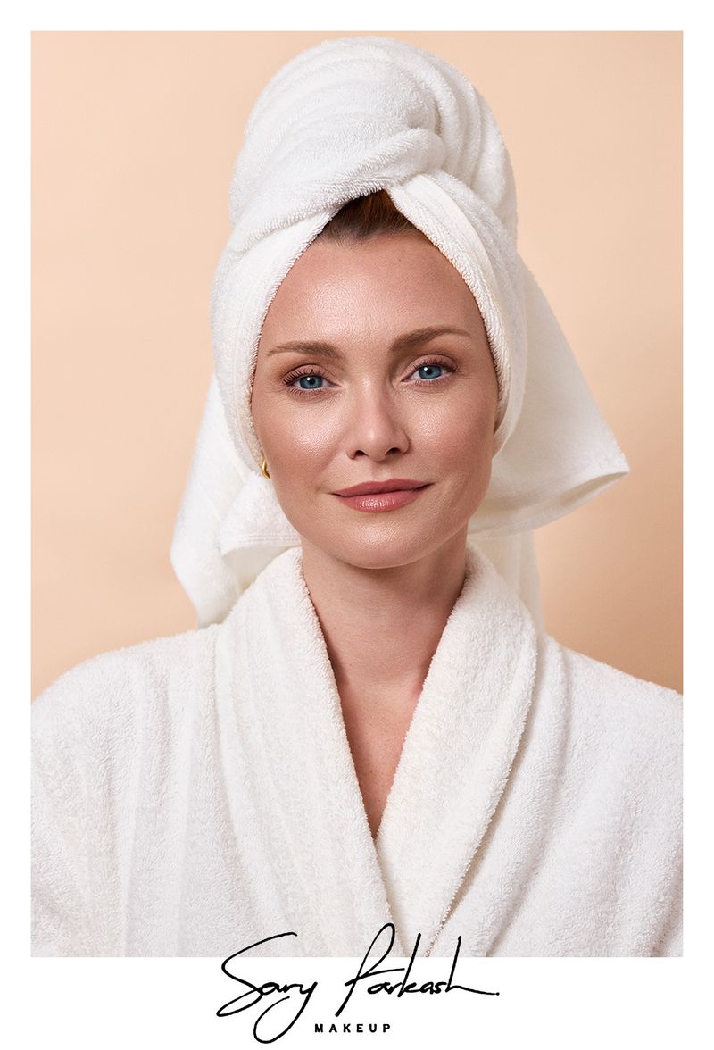 Woman in a white bathrobe and matching towel wrap on her head looking towards the camera against a neutral solid background.