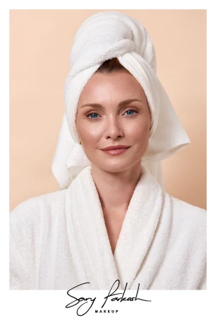 Woman in a white bathrobe and matching towel wrap on her head looking towards the camera against a neutral solid background.