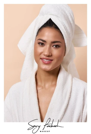 Portrait of a smiling woman with a white towel wrapped around her head and wearing a white bathrobe against a neutral background.