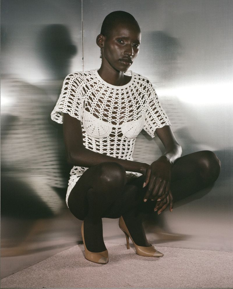 Woman in a white crocheted dress and beige high heels crouches in front of a metallic background in a studio. Fashion shot.