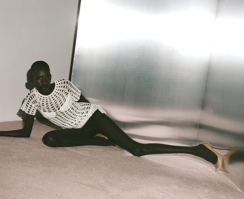 Fashion shot of an adult Black female model in a white crochet dress and nude heels, reclining on a beige floor in a studio with a metal wall.