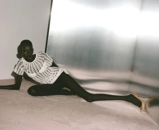 Model in white crochet top and black tights reclining against a reflective metallic panel in a studio setting