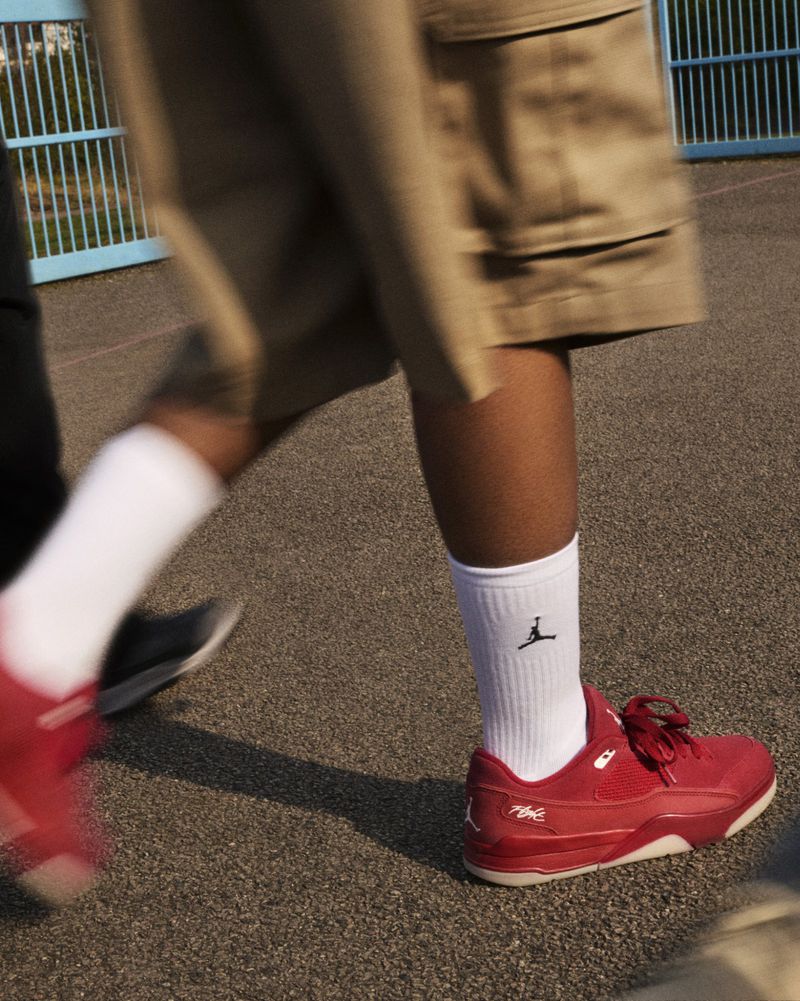 Close-up of person walking on street wearing red sneakers, white socks with a logo, and beige shorts, with another person blurred.
