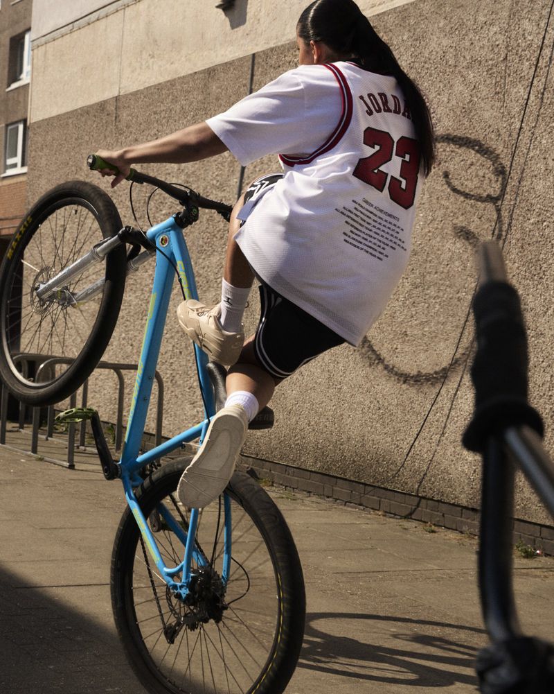 Person riding a blue BMX bike performing a wheelie on a city street in a white Jordan jersey and black shorts with a building backdrop.