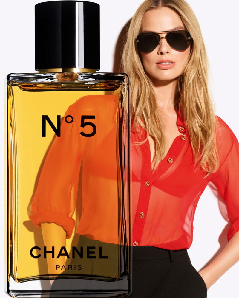 Margot Robbie poses in front of a Chanel No. 5 perfume bottle. She wears sunglasses and a red sheer blouse, showcasing fashion and beauty.