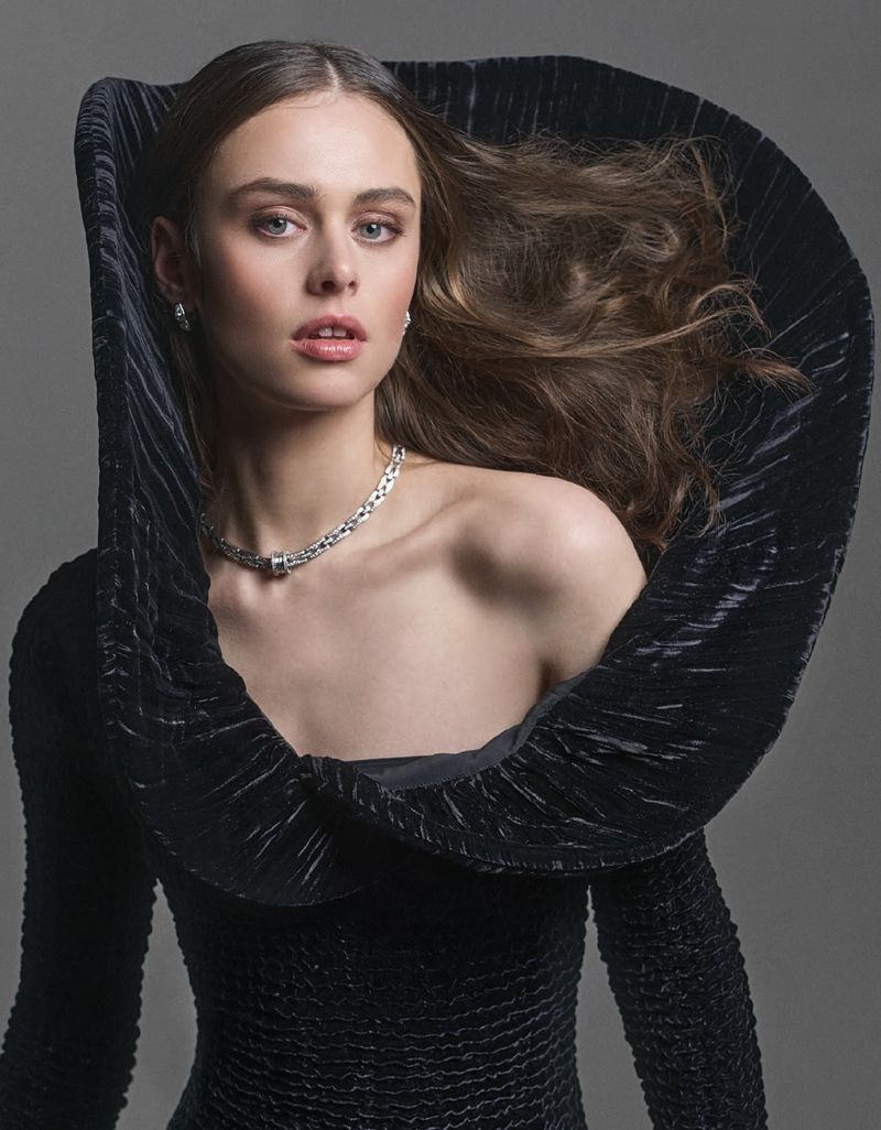 Portrait of a woman with long wavy brown hair wearing a black dress with a dramatic pleated collar and a silver chain necklace.