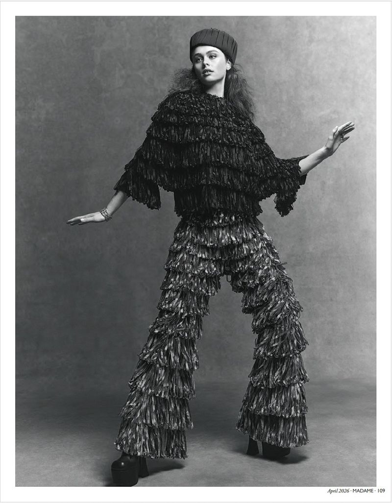 Monochrome fashion shot: a young woman poses in a fringed top and pants with her arms outstretched in a gray studio setting.
