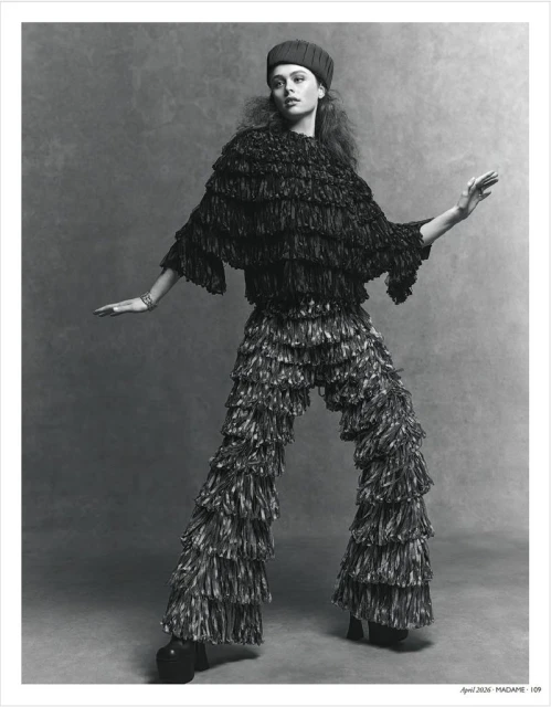 Monochrome fashion shot: a young woman poses in a fringed top and pants with her arms outstretched in a gray studio setting.