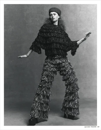 Monochrome fashion shot: a young woman poses in a fringed top and pants with her arms outstretched in a gray studio setting.