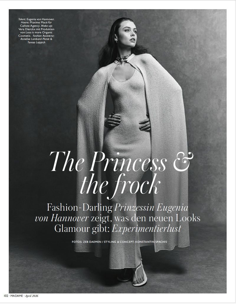 A fashion editorial shot featuring a woman wearing a long cape dress and statement jewelry, posed against a neutral grey background.