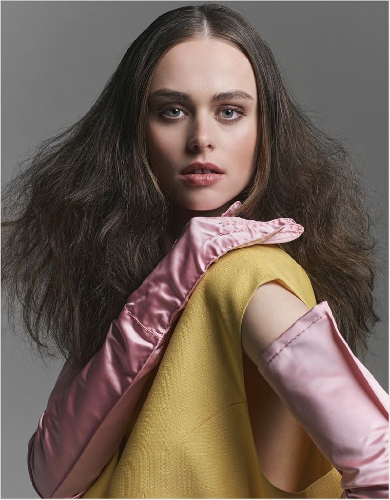 Portrait of a young woman with medium-length brown hair, wearing a mustard-yellow top and light pink satin gloves against a grey backdrop.