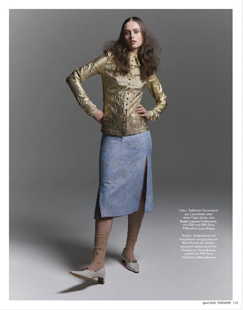 Fashion editorial shot of a woman with long brown hair in a gold button-down shirt and light blue skirt on a grey background.