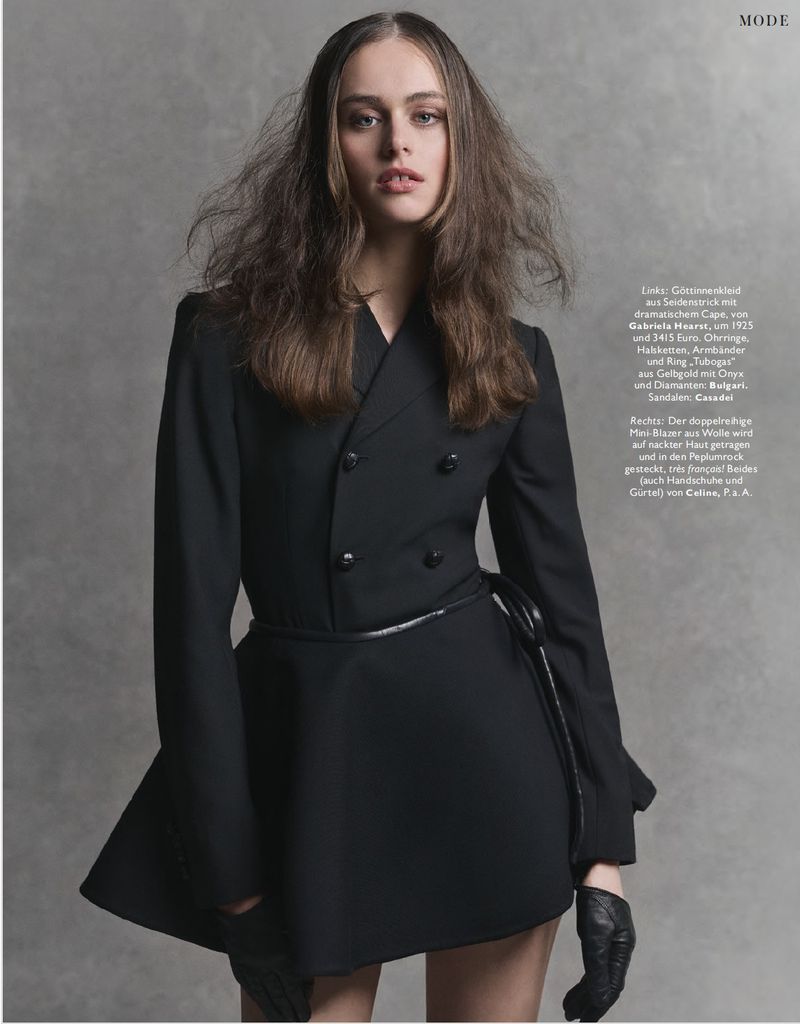 Model in black double-breasted jacket and leather gloves against a grey wall background