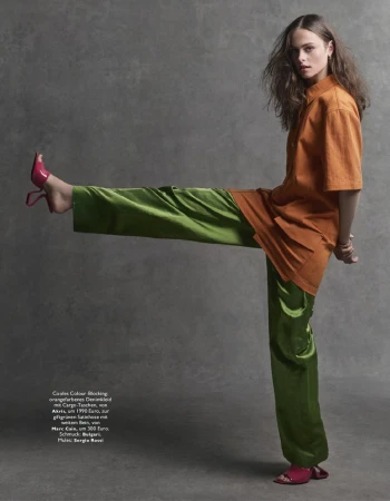 Fashionable woman in orange shirt and green pants strikes a pose against a grey backdrop. Pink high heels add a pop of color.
