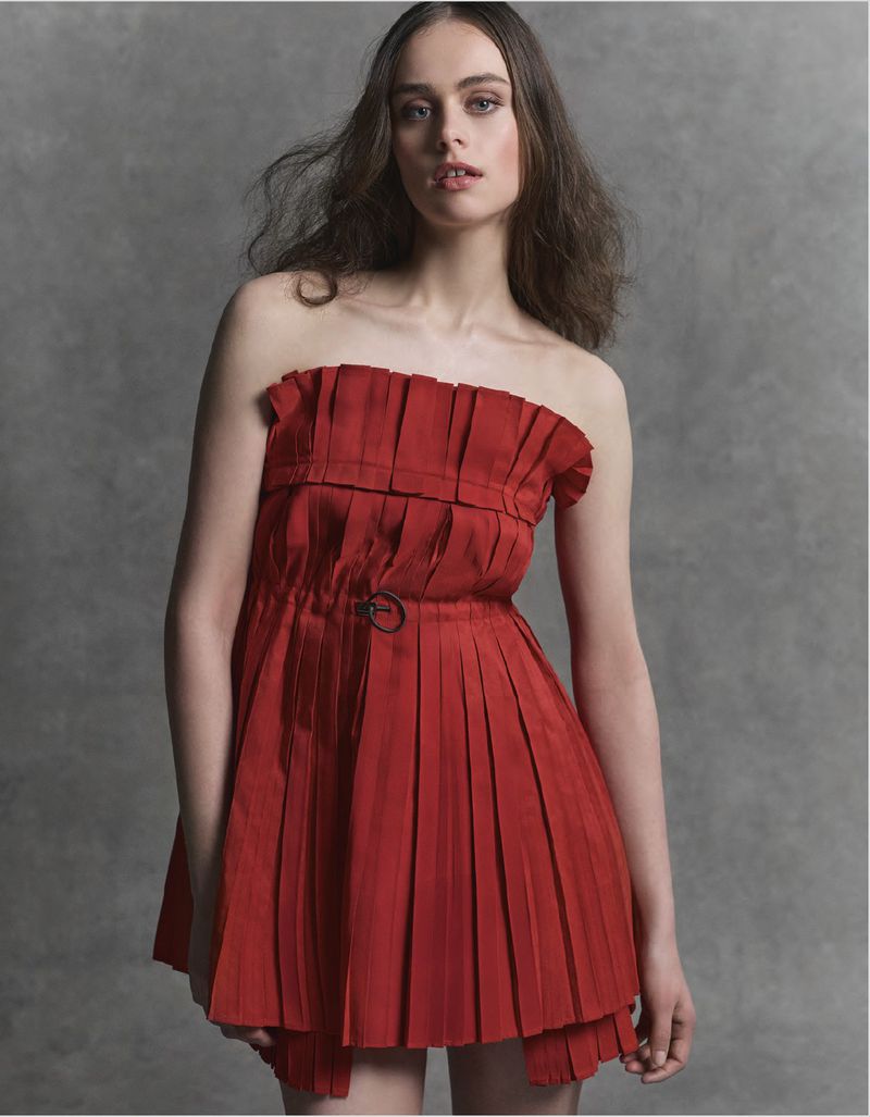 A fashionable young woman with long hair poses in a studio wearing a red strapless dress with a pleated skirt against a grey backdrop.