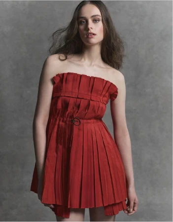 A fashionable young woman with long hair poses in a studio wearing a red strapless dress with a pleated skirt against a grey backdrop.