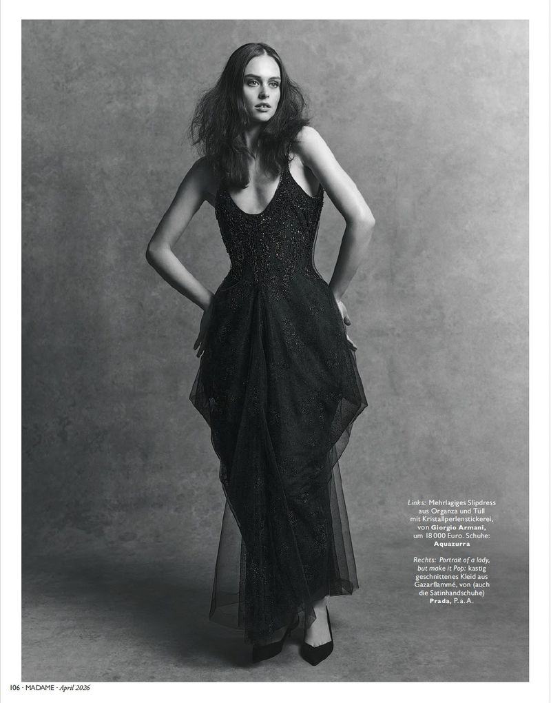 A long-haired woman in a full-length black gown poses against a grey backdrop, hands on hips, in a studio fashion shot.