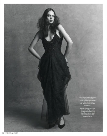 A long-haired woman in a full-length black gown poses against a grey backdrop, hands on hips, in a studio fashion shot.
