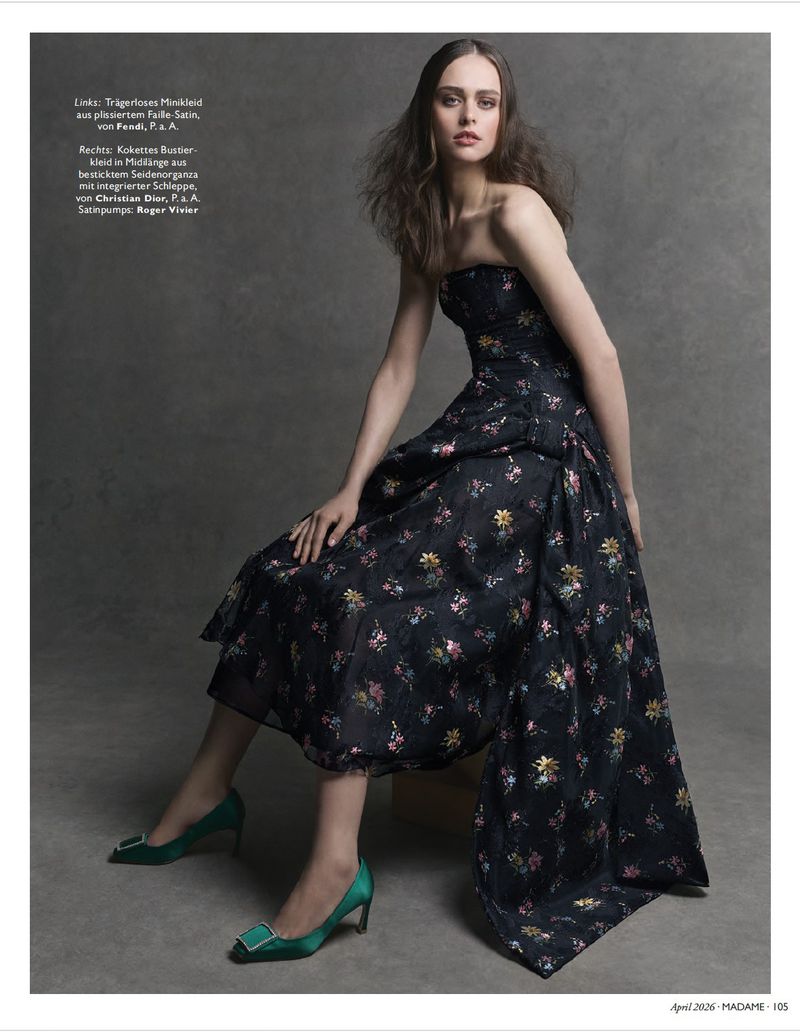 A woman poses in a strapless floral dress and green high heels in a fashion editorial, set against a neutral grey background.