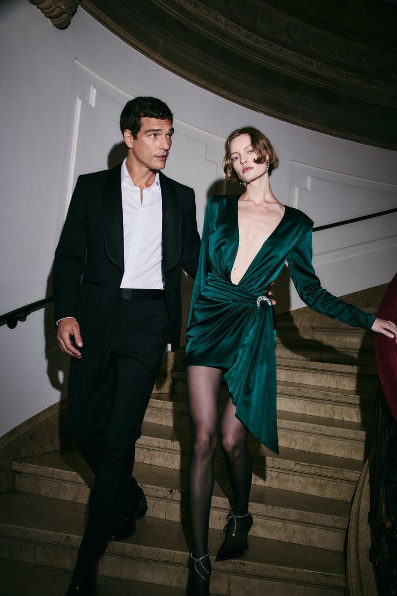 Elegant couple descends stairs: man in tuxedo, woman in emerald dress with hosiery and heels, creating a sophisticated fashion scene.
