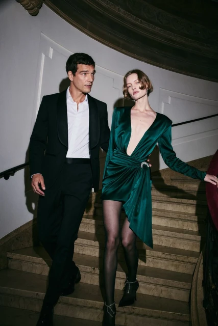 Elegant couple descends stairs: man in tuxedo, woman in emerald dress with hosiery and heels, creating a sophisticated fashion scene.