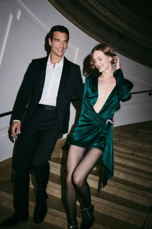 Man in a black tuxedo and a woman in a green silk dress descending a grand indoor staircase together.