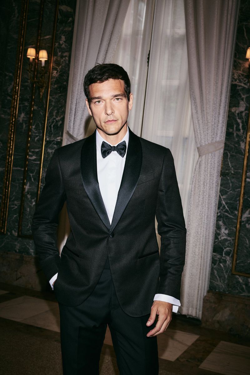 Portrait of an adult male in a black tuxedo with a bow tie against a neutral background with wall paneling and sheer curtain.