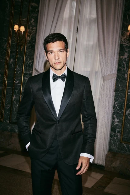Portrait of an adult male in a black tuxedo with a bow tie against a neutral background with wall paneling and sheer curtain.