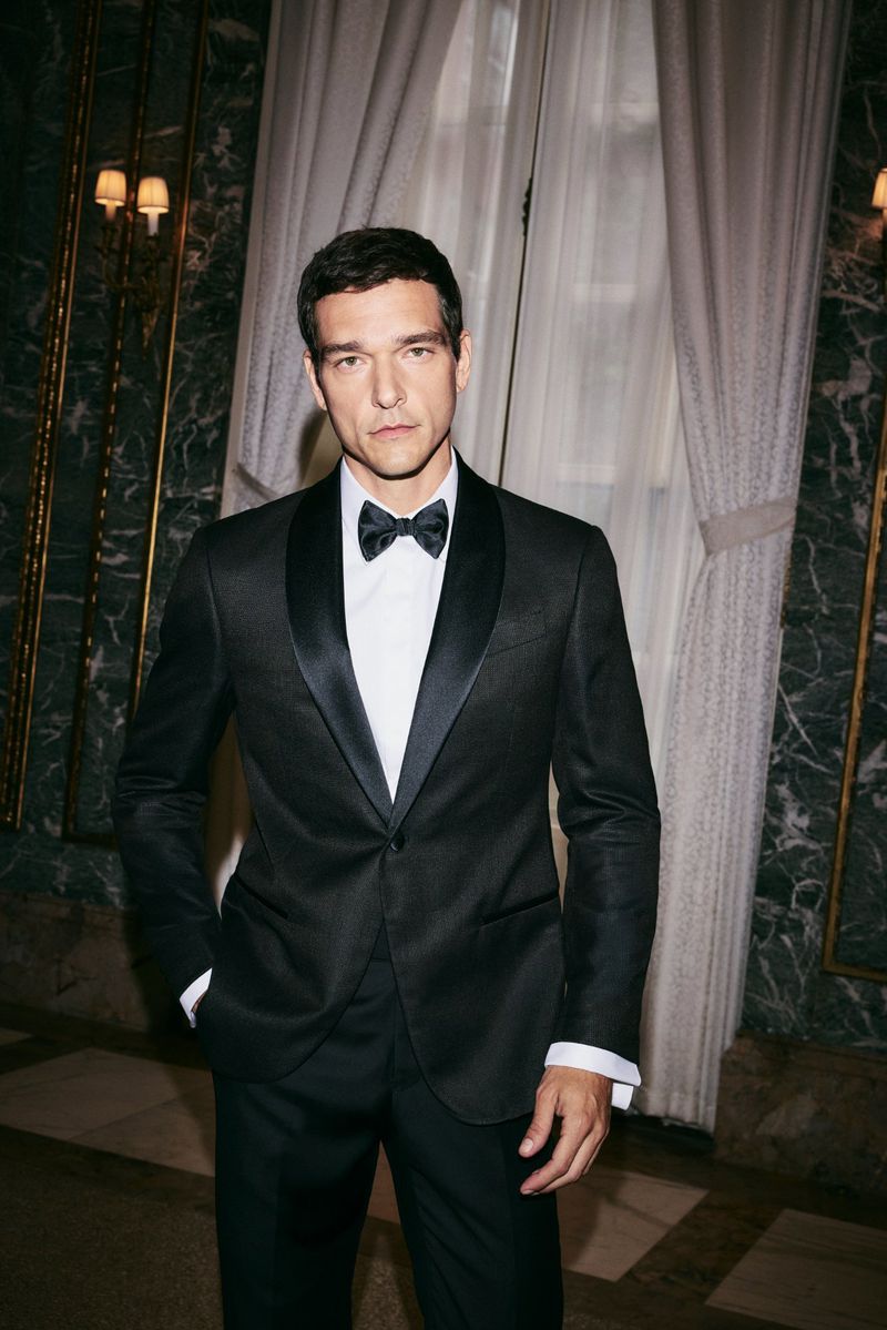 Elegant man in a black tuxedo with a bow tie stands in a marble-walled interior with sconces and sheer curtains creating a classic aesthetic.